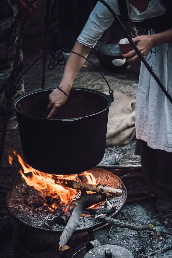 A cauldron over a open fire with a old fashioned dressed woman stirring the pot