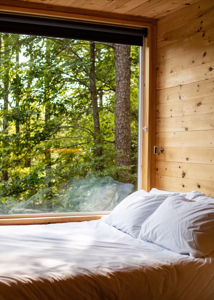 Looking out of the window onto a forest scene from a wooden cabin and made bed with white cover and pillows.